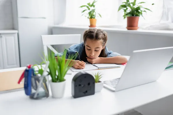 Niño escribiendo en el libro de copias cerca de la computadora portátil borrosa y plantas en casa - foto de stock