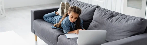 Preteen kid looking at notebook near laptop on couch at home, banner — Stock Photo