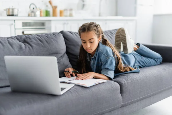 Kid holding pen near notebook and blurred laptop on couch — Stock Photo