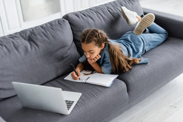 Preteen kid writing on notebook near laptop on couch — Stock Photo