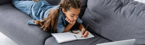 Colegiala escribiendo en el cuaderno cerca de la computadora portátil en el sofá en casa, pancarta - foto de stock