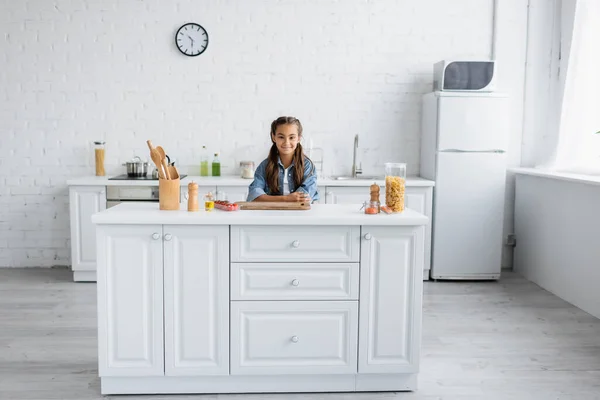 Lächelndes Kind blickt in die Kamera neben Makkaroni und Kirschtomaten in der Küche — Stockfoto