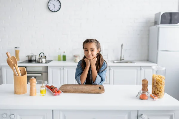 Bambino sorridente che guarda la fotocamera vicino al tagliere e agli ingredienti in cucina — Foto stock