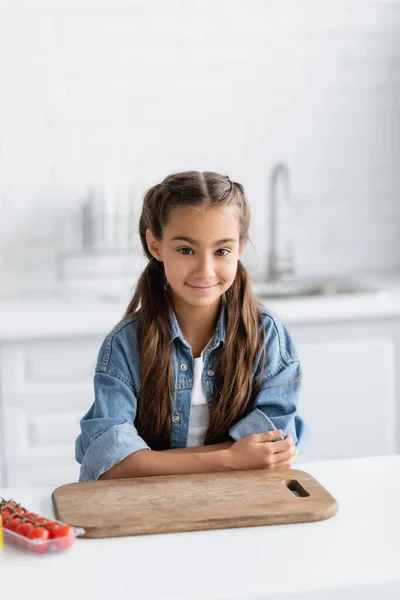 Улыбающийся ребенок смотрит в камеру рядом с разделочной доской и помидорами черри на кухне — стоковое фото
