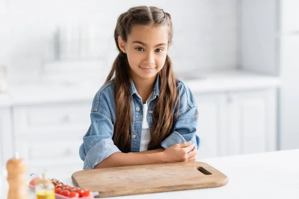 Criança pré-adolescente olhando para a câmera perto de tábua de corte e tomates cereja borrados na cozinha — Fotografia de Stock