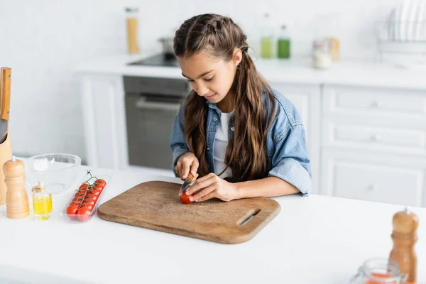 Ребенок режет помидор черри возле оливкового масла на кухне — стоковое фото