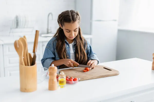 Criança pré-adolescente cortando tomate cereja perto de utensílio de cozinha e azeite na cozinha — Fotografia de Stock