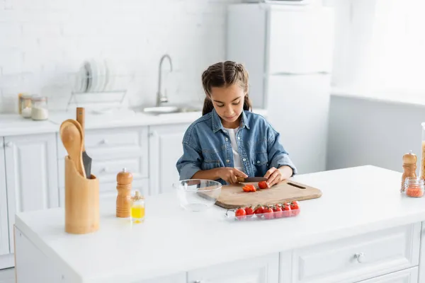 Preteen fille coupe tomate cerise près de l'huile d'olive et bol dans la cuisine — Photo de stock