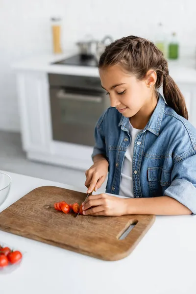 Enfant souriant coupant la tomate cerise sur planche à découper en bois dans la cuisine — Photo de stock