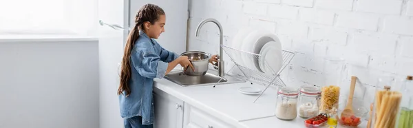 Side view of child pouring water in saucepan near food in kitchen, banner — Stock Photo