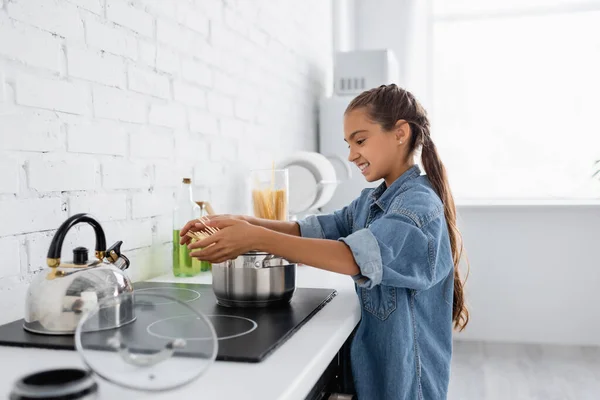 Side view of cheerful girl holding macaroni near saucepan and kettle on stove in kitchen — Stock Photo