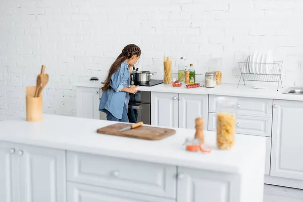 Малюк стоїть біля каструлі на плиті та інгредієнтів на кухні — стокове фото