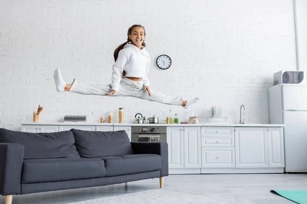 Cheerful kid doing split while jumping near couch in kitchen — Stock Photo