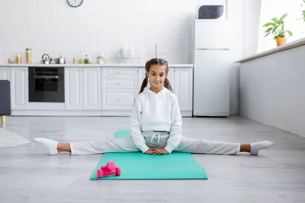 Preteen girl looking at camera while doing split near dumbbells in kitchen — Stock Photo