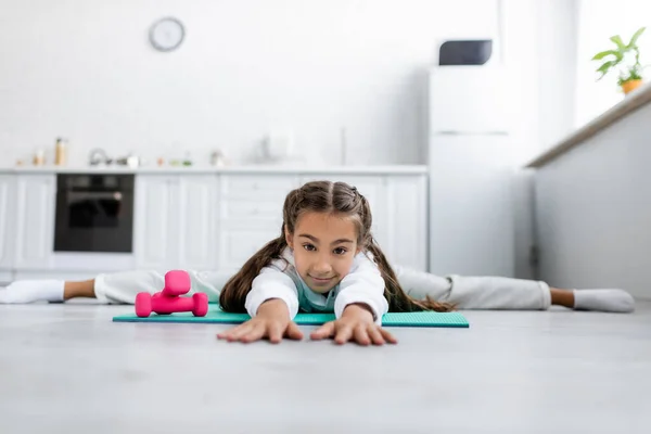 Surface level of child looking at camera while doing split near dumbbells in kitchen — Stock Photo