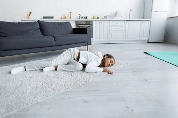 Sonriente niña preadolescente acostada en la alfombra cerca de la alfombra de fitness en la cocina - foto de stock