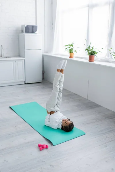 Happy kid training on fitness mat near dumbbells in kitchen — Stock Photo