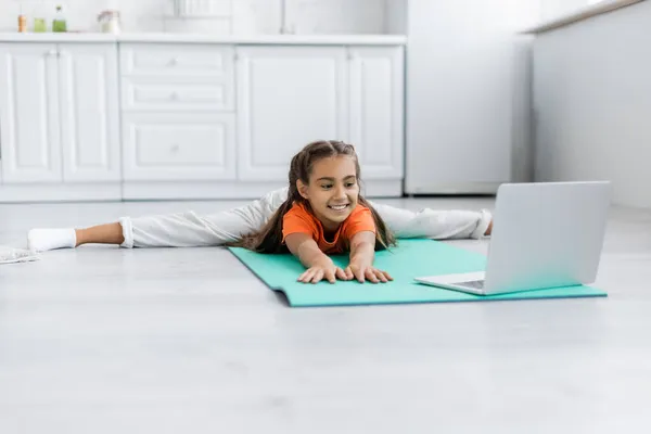 Menina alegre que se estende perto do laptop no tapete de fitness em casa — Fotografia de Stock