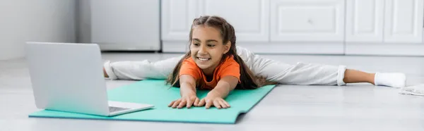 Criança sorrindo fazendo split perto de laptop no tapete de fitness em casa, banner — Fotografia de Stock