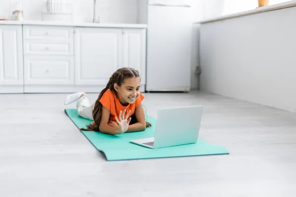 Lächelnd preteen girl having video call on laptop on fitness matte at home — Stockfoto