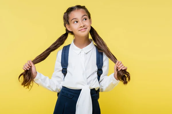 Dreamy schoolgirl with backpack touching hair isolated on yellow — Stock Photo