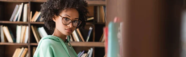 Joven afroamericana mujer en gafas mirando la cámara cerca de estantería, pancarta - foto de stock