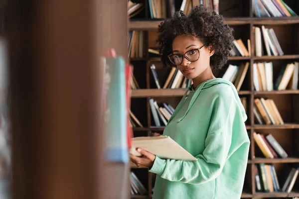 Young african american woman in eyeglasses looking at camera while holding book near bookshelf — Stock Photo