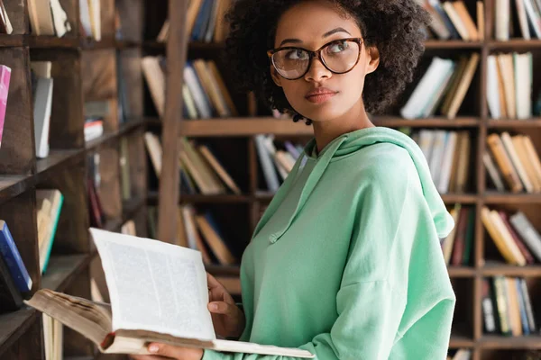 Jovem afro-americana em óculos segurando livro perto estante — Fotografia de Stock