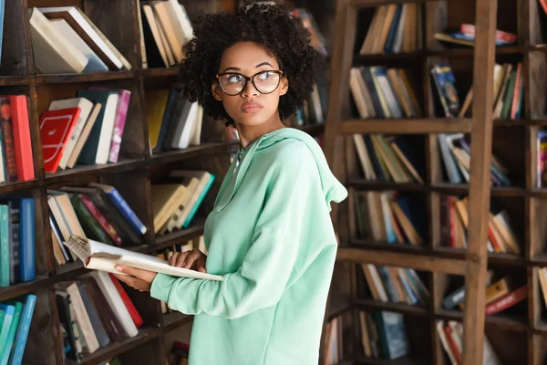 Jovem estudante afro-americano em óculos segurando livro perto estante — Fotografia de Stock