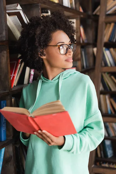 Jovem estudante afro-americano em óculos segurando livro enquanto olha para longe perto estante — Fotografia de Stock