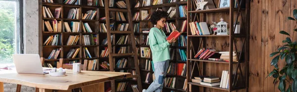 Joven afroamericano estudiante en gafas lectura libro cerca de gadgets en escritorio, bandera - foto de stock