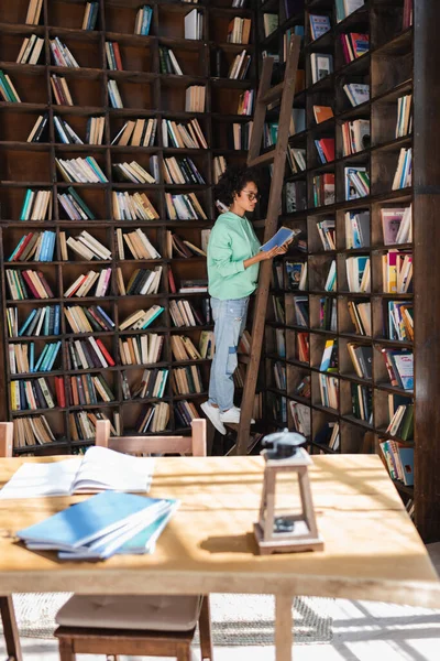 Estudiante afroamericano en gafas de pie en escalera de madera mientras lee libro cerca de cuadernos borrosos en el escritorio - foto de stock