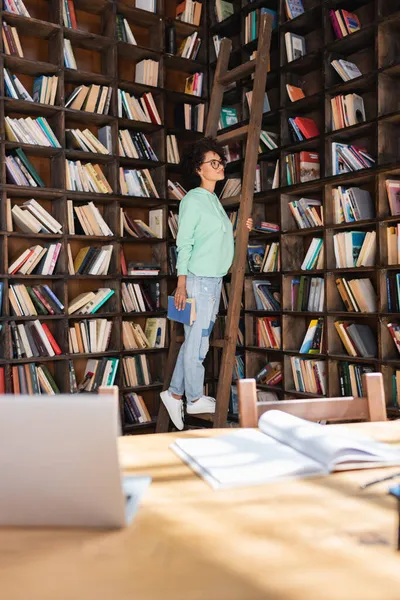 Young african american student in eyeglasses standing on wooden ladder and holding book — Stock Photo
