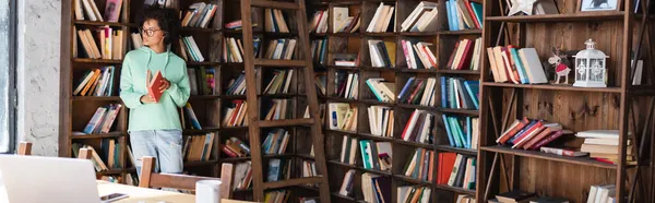 Jeune étudiant afro-américain en lunettes tenant le livre près de l'échelle en bois et des dispositifs sur le bureau, bannière — Photo de stock