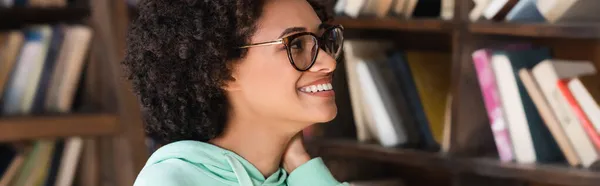 Happy african american student in eyeglasses looking at books on bookcase, banner — Stock Photo
