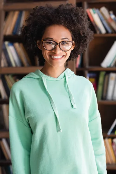 Веселий афроамериканський студент в окулярах дивиться на камеру біля книжкової полиці — стокове фото