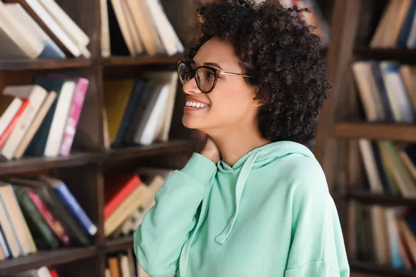 Cheerful african american student in eyeglasses looking at bookshelf — Stock Photo