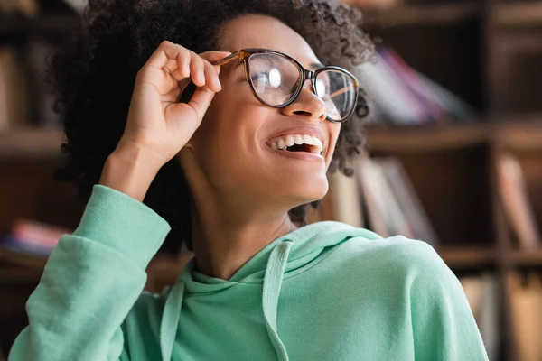 Positive african american student adjusting eyeglasses and looking away — Stock Photo