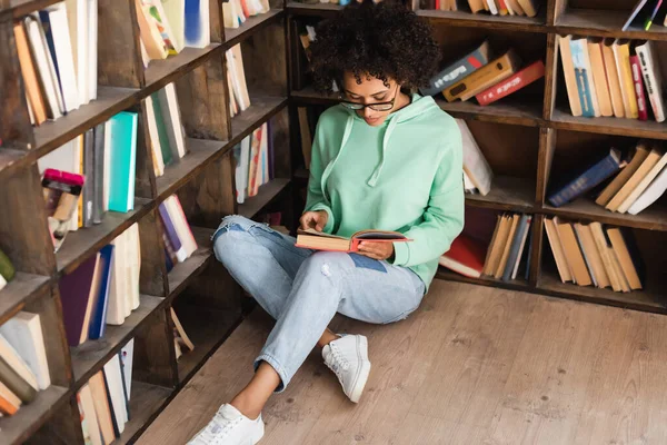 Curly african american student in eyeglasses sitting on floor and reading book in library — Stock Photo