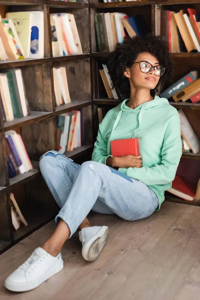 Молодая африканская студентка в очках сидит на полу с книгой возле книжной полки в библиотеке — стоковое фото