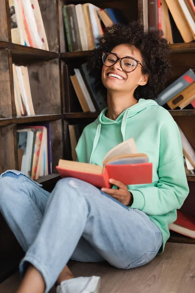 Estudiante afroamericano feliz en anteojos sentados en el piso con libro en la biblioteca - foto de stock