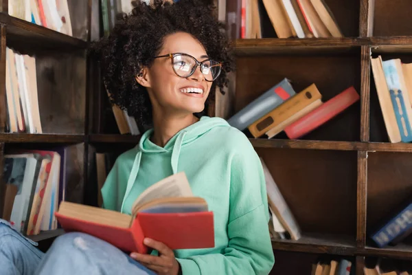 Estudiante afroamericano feliz en anteojos sentado con libro en la biblioteca - foto de stock