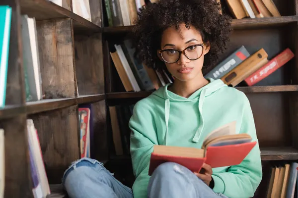 Bouclé afro-américain étudiant en lunettes tenant livre à la bibliothèque — Photo de stock