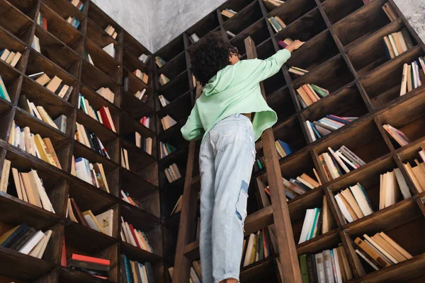 Low angle view of young african american student standing on wooden ladder in library — Stock Photo