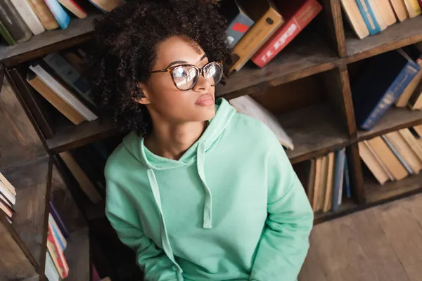 Aus der Vogelperspektive: junge afrikanisch-amerikanische Studentin mit Brille schaut in Bibliothek weg — Stockfoto