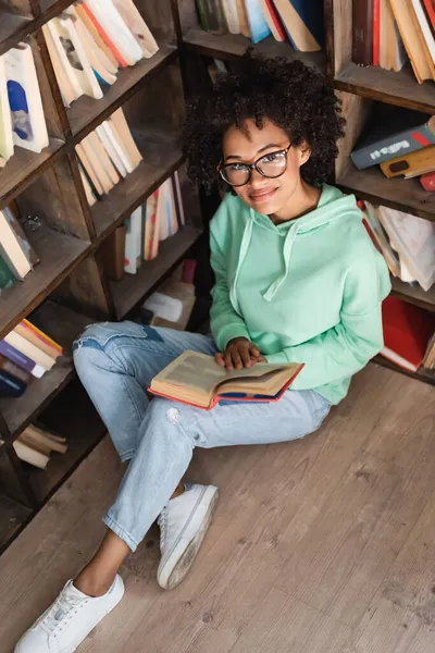 Hochwinkelaufnahme eines lächelnden afrikanisch-amerikanischen Studenten mit Brille, der mit Buch in der Bibliothek sitzt — Stockfoto