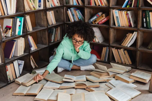 Inteligente estudante afro-americano em óculos sentado cercado por livros em biblioteca — Fotografia de Stock