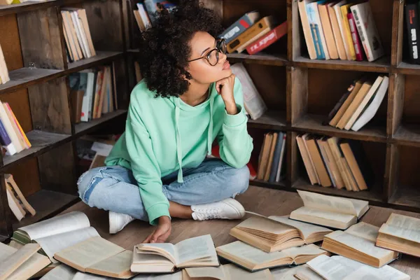 Pensativo afroamericano estudiante en gafas sentado rodeado de libros en la biblioteca - foto de stock