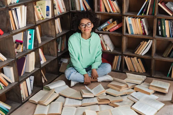 Zufrieden afrikanisch-amerikanischer Student mit Brille sitzt mit überkreuzten Beinen umgeben von Büchern in der Bibliothek — Stockfoto