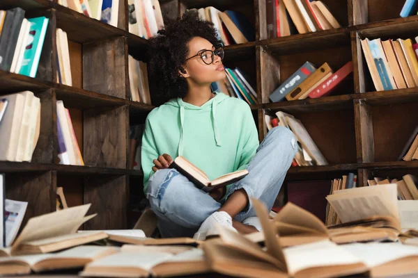 Mujer afroamericana inteligente en anteojos sentada rodeada de libros en la biblioteca - foto de stock